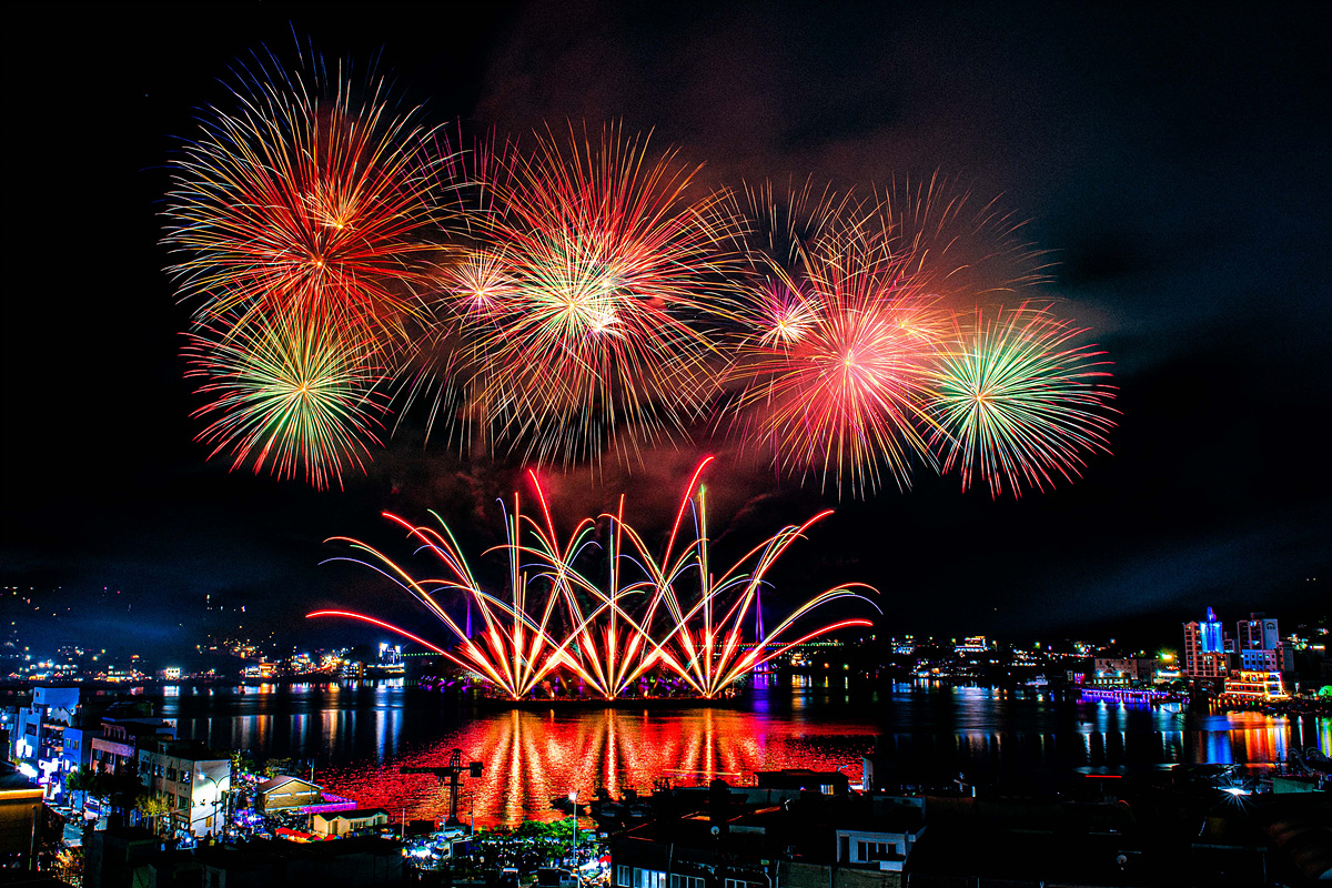 밤하늘에 화려한 불꽃놀이가 펼쳐지고 있으며, 불꽃이 반사된 물결이 아름답게 빛나고 있다. 주변 건물과 조명이 어우러져 축제 분위기를 자아내고 있다.