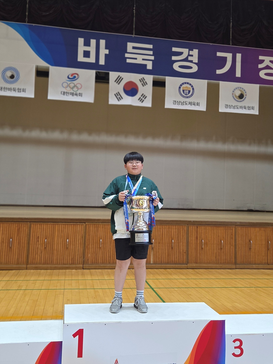 한 어린이가 바둑 대회에서 1등 트로피를 들고 시상대에 서 있다. 배경에는 바둑 관련 배너가 걸려 있다.