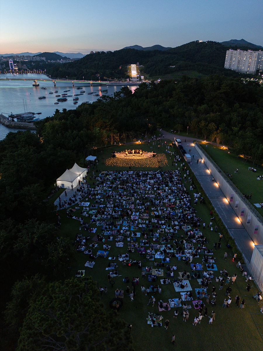 여수 밤바다를 배경으로 넓은 잔디밭 위에서 진행된 ‘캔들라이트 콘서트’ 전경. 관람객들이 돗자리를 펴고 앉아 공연을 관람하고 있으며, 무대 위에는 수많은 촛불이 은은하게 빛나고 있다