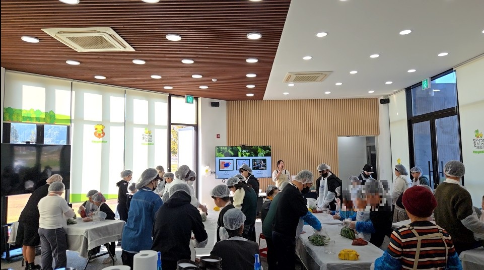 여수 드림스타트 아동과 가족이 돌산갓 체험장 실내에서 갓김치 만들기와 고추장 담그기 등 식재료 활용 영양교육을 체험하고 있다. 모든 참가자가 위생 모자와 장갑을 착용한 채 강사의 설명을 들으며 조리 과정을 진행하는 모습