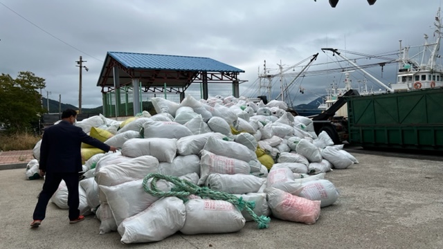 항구에 쌓여 있는 대량의 인양쓰레기 마대 더미를 한 남성이 살펴보고 있는 모습. 어선들이 정박한 선착장 배경으로 흰색·회색 마대 봉투가 높게 쌓여 있다.