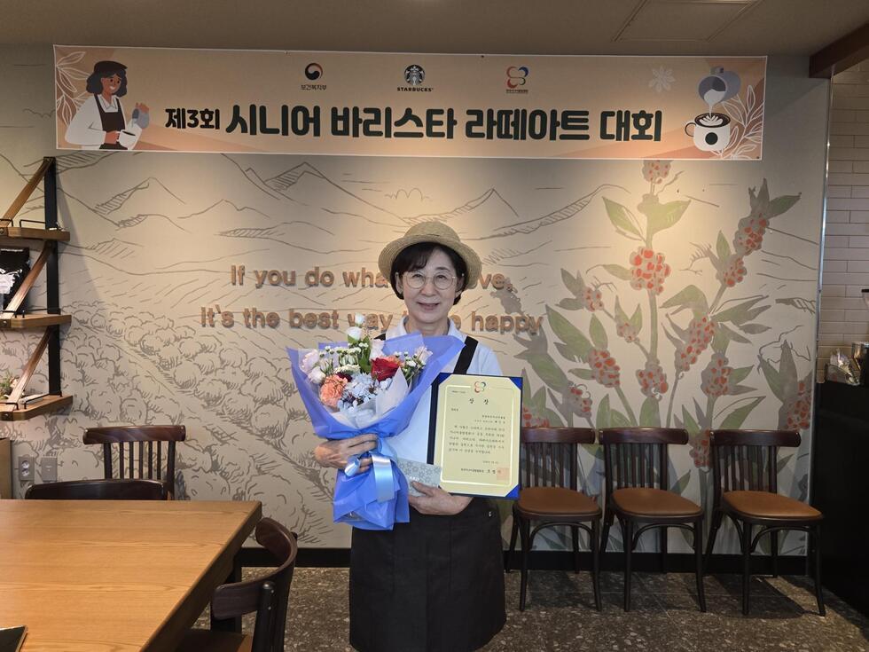 시니어 바리스타 대회에서 꽃다발과 상장을 들고 있는 참가자가 기념 촬영을 하고 있다. 배경에는 대회 관련 현수막과 카페 인테리어가 보인다.