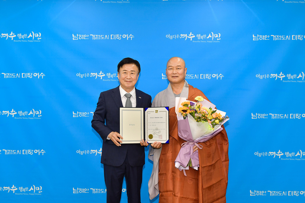 여수에서 한 남성과 스님이 상장과 꽃다발을 들고 기념사진을 찍고 있다. 배경에는 여수의 슬로건이 적힌 파란색 배너가 있다.