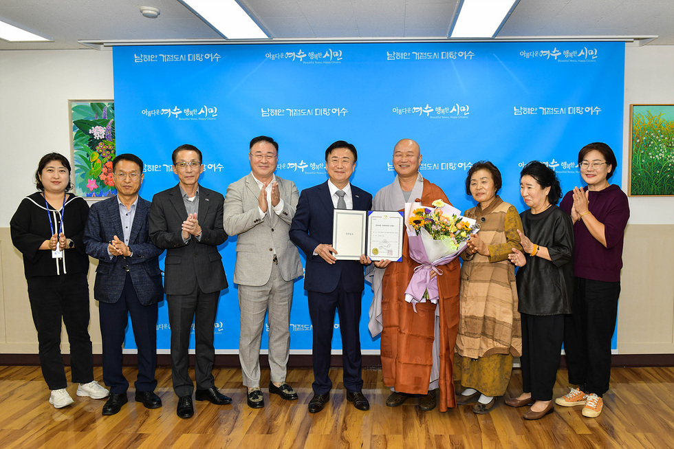 여수에서 여러 사람들이 모여 기념사진을 찍고 있으며, 한 남성이 상장과 꽃다발을 들고 있다. 배경에는 여수의 슬로건이 적힌 현수막이 있다.