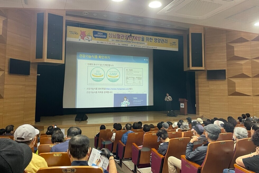 2024 시니어 건강관 행사에서 강사가 건강기능식품에 대해 설명하고 있으며, 청중이 강의를 듣고 있는 모습이다.