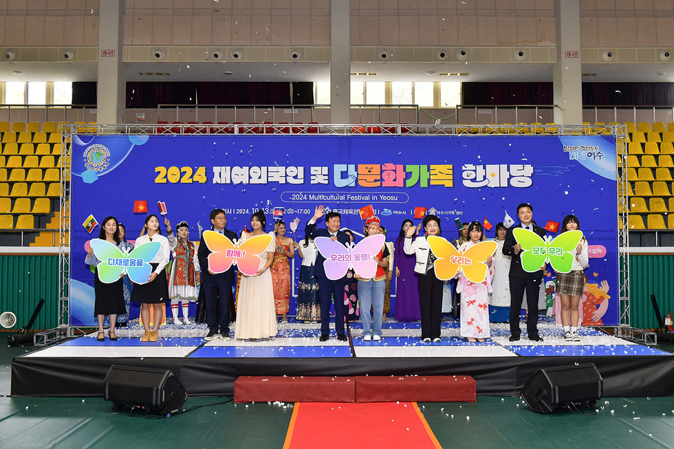 2024 재여외국인 및 다문화가족 한마당 행사에서 여러 사람들이 각기 다른 색상의 나비 모양의 표지를 들고 서 있다. 배경에는 행사 정보가 적힌 현수막이 걸려 있다.