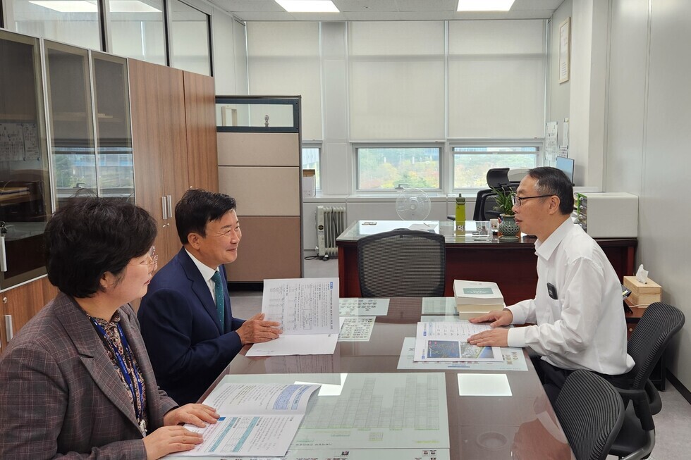 세 명의 남녀가 사무실에서 회의 중이며, 각자 문서를 들고 대화하고 있다. 배경에는 사무실 가구와 창문이 보인다.