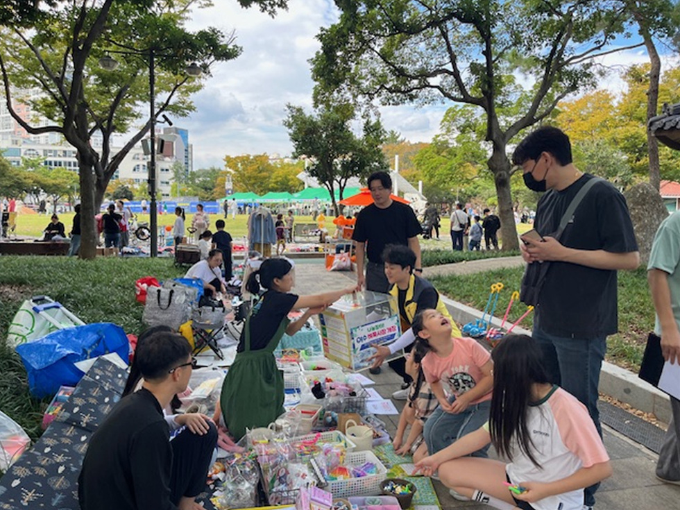 공원에서 여러 사람이 모여 다양한 물건을 판매하고 있는 장면이다. 한 여성이 물건을 손님에게 보여주고 있으며, 주변에는 아이들과 다른 사람들이 있다.