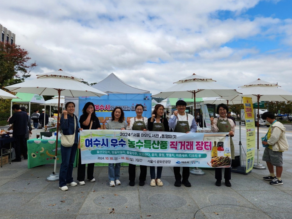 여수시 우수 농수산물 직거래 장터에서 사람들이 홍보 배너를 들고 포즈를 취하고 있다. 행사 날짜는 2024년 10월 18일부터 20일까지이다.