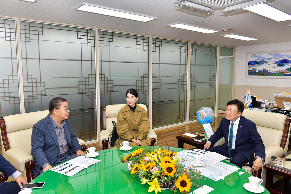 회의실에서 세 명의 사람들이 앉아 대화하고 있으며, 테이블 위에는 꽃다발과 여러 자료가 놓여 있다.