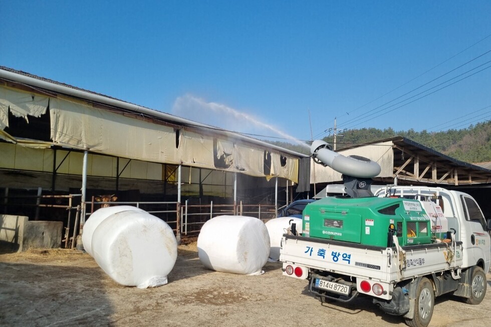 농장에서 가축 방역을 위해 소독기가 작동되고 있는 모습으로, 소독기가 건물 위로 물을 분사하고 있다.