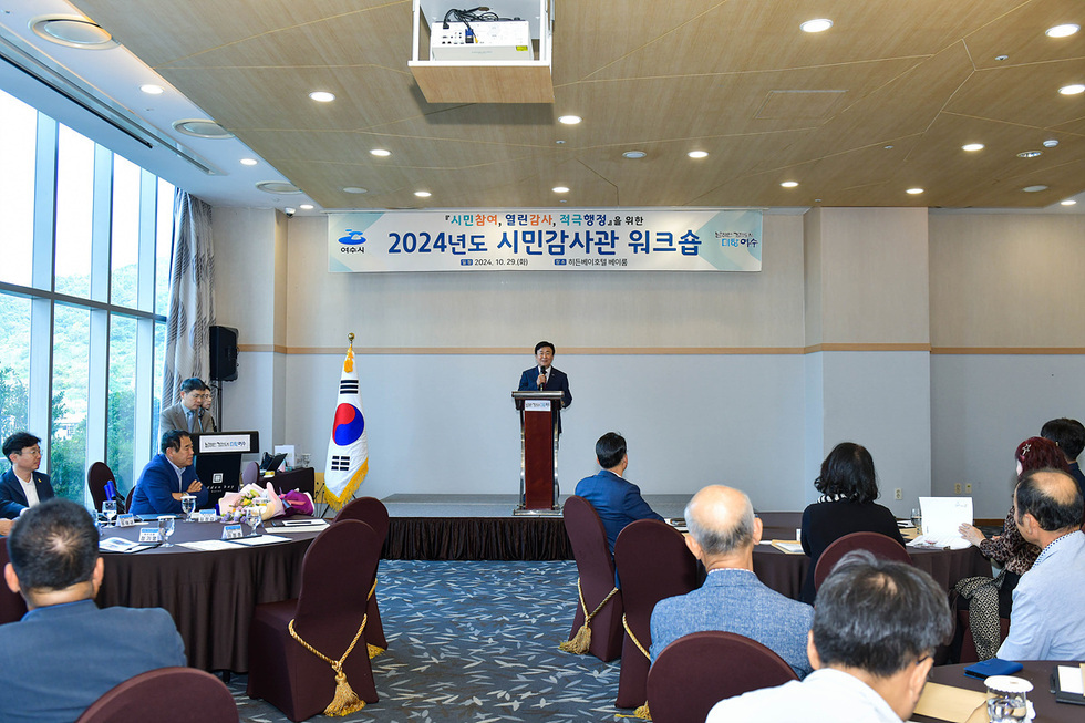 여수시에서 2024년도 시민감사관 워크숍이 진행되고 있으며, 한 연사가 무대에서 발표하고 있다. 참석자들이 테이블에 앉아 경청하고 있다.