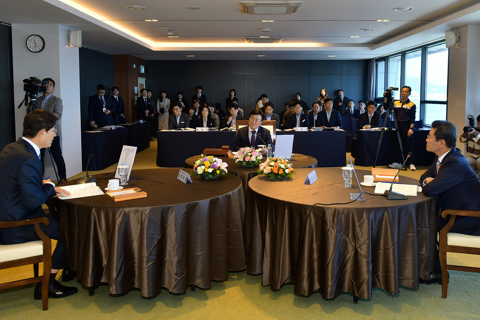 회의 중인 여러 사람들로 구성된 장면으로, 두 개의 원형 테이블에 앉은 사람들이 대화하고 있으며, 배경에는 청중이 앉아 있다.