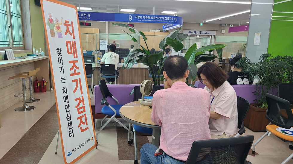 여수시치매안심센터에서 한 남성이 직원과 상담을 하고 있으며, 배경에는 치매조기검진 안내 현수막이 보인다.