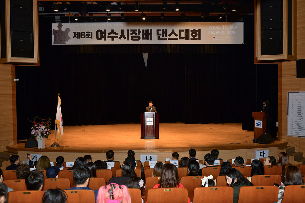 여수문화예술회관에서 제6회 여수시장배 댄스대회가 열리고 있으며, 무대에서 한 남성이 발표를 하고 있다. 관객들이 앉아 대회를 관람하고 있다.