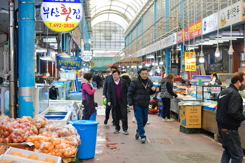 부산의 전통 시장에서 사람들이 쇼핑을 하고 있으며, 다양한 해산물과 과일이 진열된 상점들이 보인다.