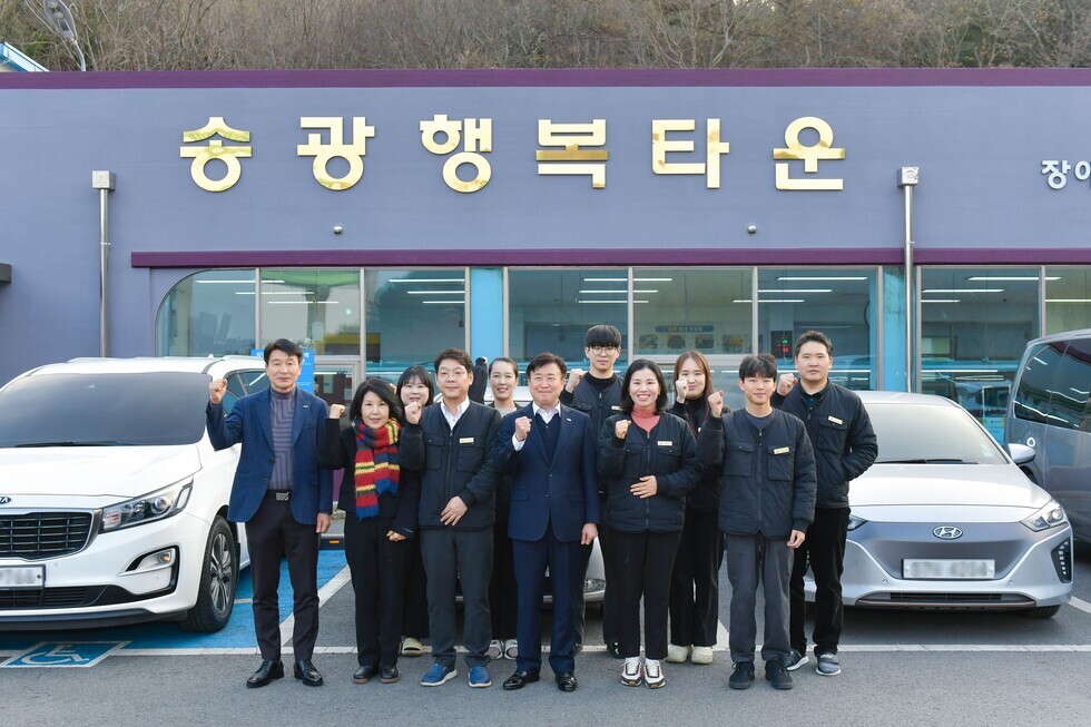 송광 행복타운 앞에서 직원들이 모여 포즈를 취하고 있다. 배경에는 두 대의 차량과 건물이 보인다.