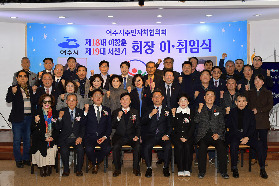 여수시 주민자치협의회에서 제18대 이창훈과 제19대 서선기의 회장 취임식이 진행되고 있다. 참석자들이 단체 사진을 찍으며 주먹을 쥐고 포즈를 취하고 있다.