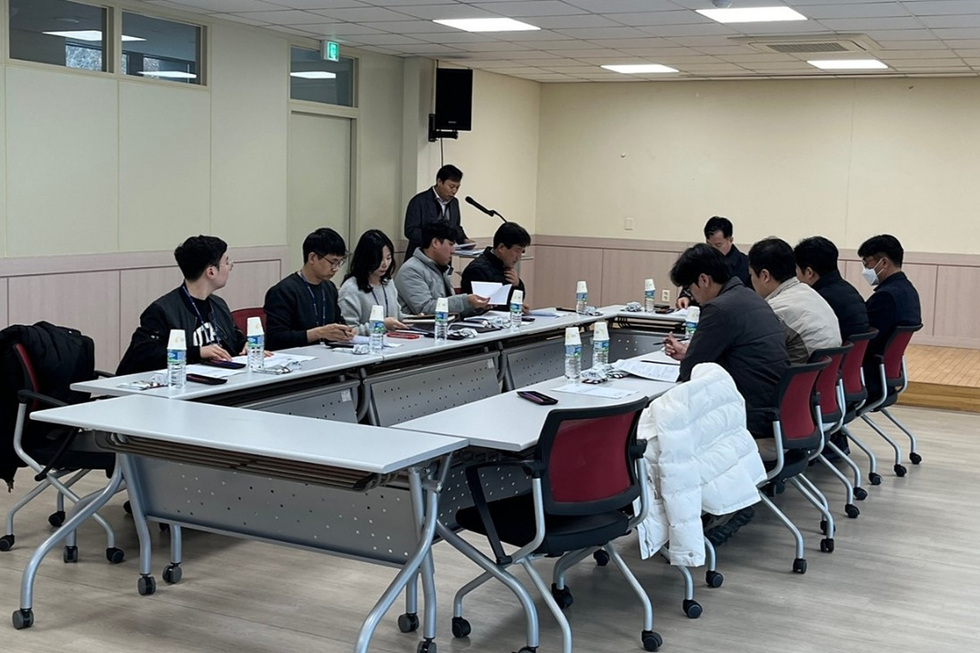 회의실에서 여러 사람이 테이블에 앉아 회의 중이다. 한 사람이 발표를 하고 있으며, 참석자들은 노트와 스마트폰을 사용하고 있다.