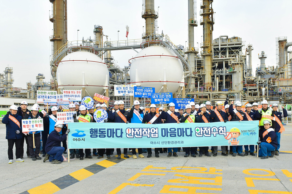 여수시에서 안전작업을 촉구하는 행사에 참여한 사람들이 안전 수칙을 담은 현수막을 들고 있다. 배경에는 산업 시설이 보인다.