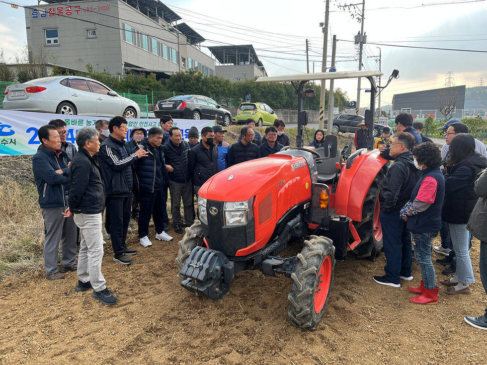 농업기계 안전교육을 받기 위해 모인 사람들 앞에 트랙터가 놓여 있다. 교육생들이 트랙터 주변에 모여 설명을 듣고 있다.