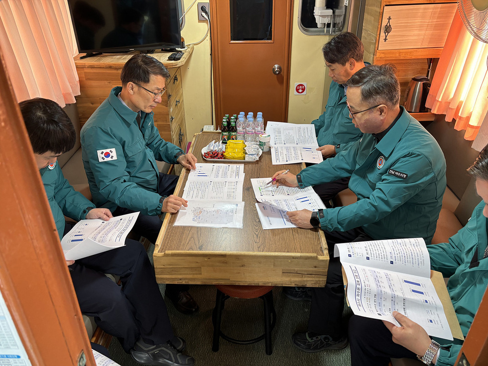 여섯 명의 남성이 회의 테이블에 앉아 자료를 검토하고 있는 모습이다. 그들은 각자 문서를 보며 토론하고 있으며, 배경에는 TV와 주방용품이 보인다.