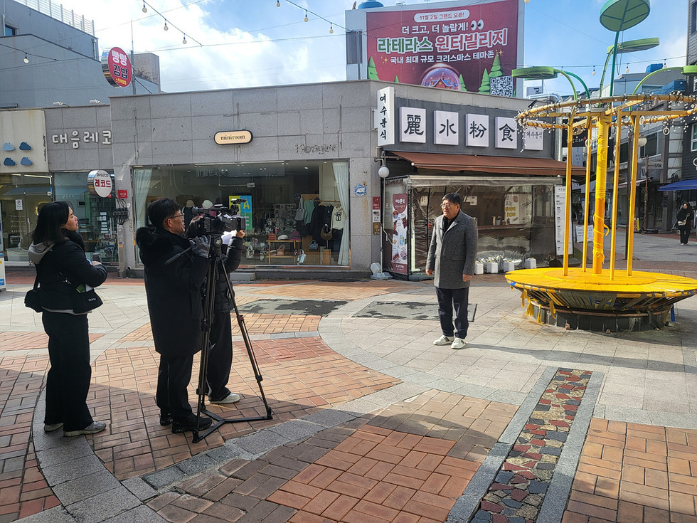 한 남성이 거리에서 촬영 중인 기자와 카메라맨 앞에 서 있다. 배경에는 크리스마스 테마축제를 홍보하는 현수막이 걸려 있다.