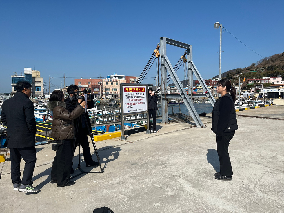 어선이 있는 항구에서 한 여성이 카메라를 향해 서 있고, 다른 사람들이 촬영을 하고 있는 장면이다. 배경에는 알림사항이 적힌 현수막이 보인다.