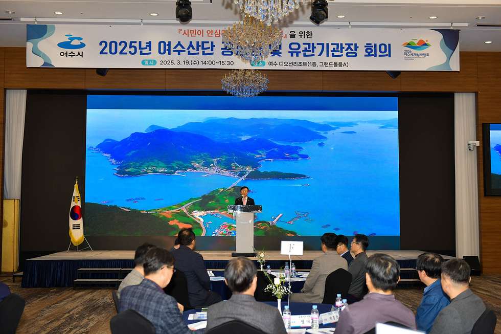 여수에서 2025년 여수산단 공공 및 유관기관장 회의가 진행되고 있으며, 발표자가 무대에서 연설하고 있다. 배경에는 여수의 해안 경관이 보인다.