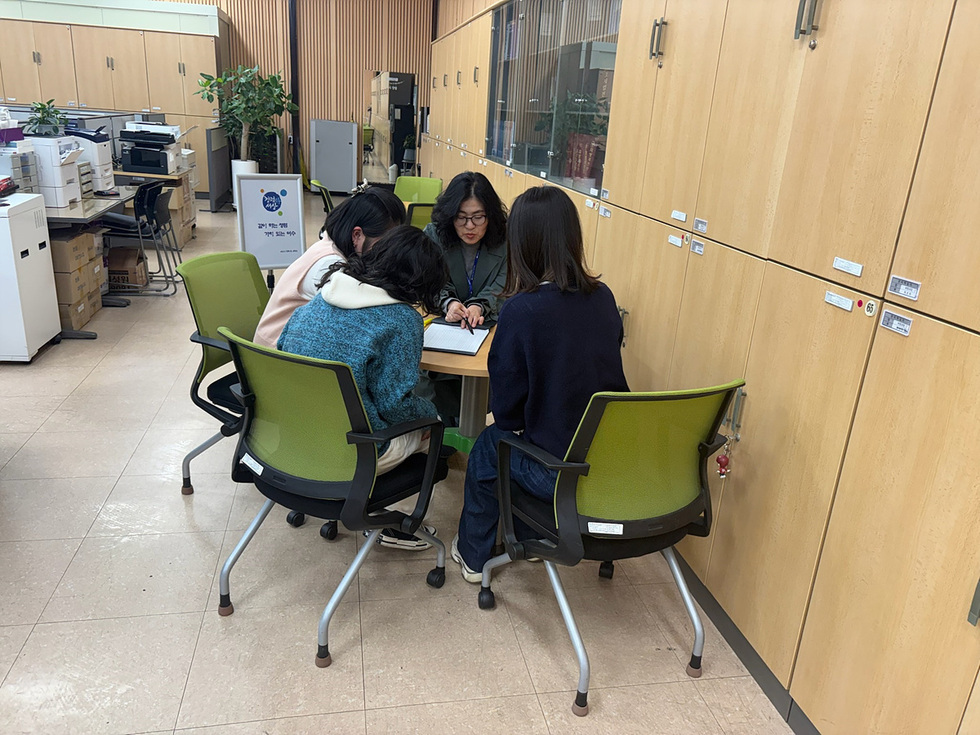 네 명의 사람들이 테이블에 앉아 함께 작업하고 있는 모습이다. 주변에는 사무기기와 캐비닛이 배치되어 있다.