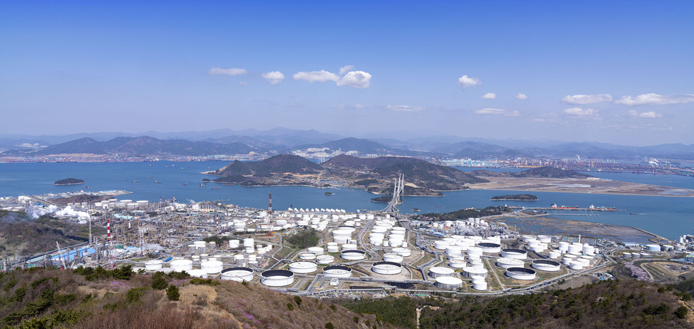 산에서 바라본 해안가의 산업 단지와 탱크들이 보이는 풍경으로, 바다와 섬, 그리고 멀리 도시의 모습이 함께 담겨 있다.
