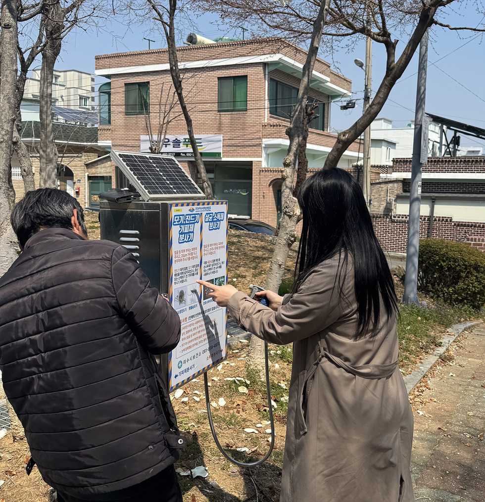 두 사람이 미기상기기 사용법 안내판을 보고 있는 장면으로, 한 사람은 안내판을 가리키고 다른 사람은 스마트폰을 들고 있다.