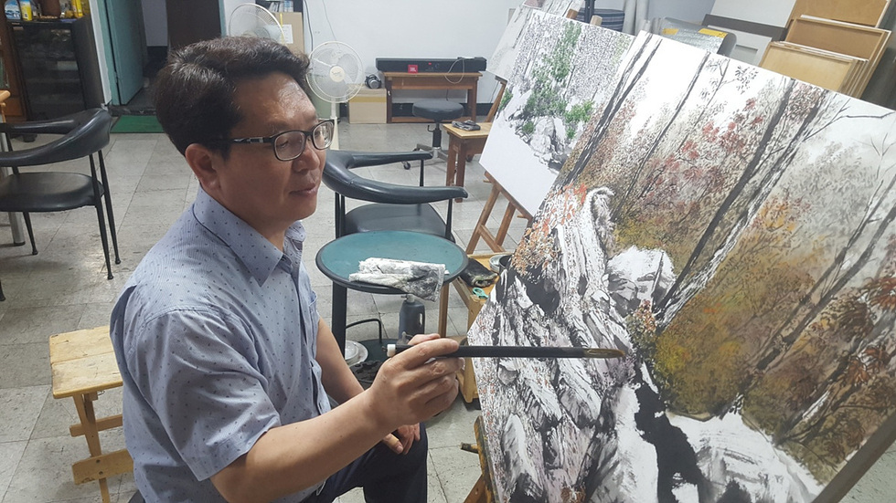 한 남성이 작업실에서 그림을 그리고 있다. 그는 붓을 들고 있으며, 배경에는 나무와 바위가 있는 풍경화가 보인다.