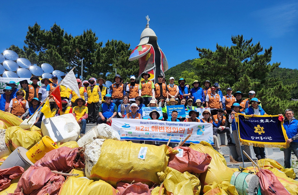 여수시에서 해변 정화 활동에 참여한 자원봉사자들이 기념사진을 찍고 있다. 배경에는 해양 쓰레기가 쌓여 있고, 여러 개의 현수막이 보인다.