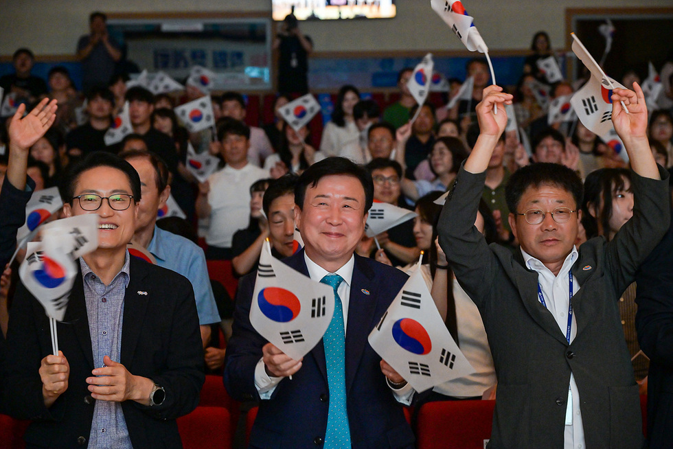 관중들이 한국 국기를 흔들며 응원하는 모습으로, 앞쪽에 세 명의 남성이 웃으며 손을 흔들고 있다.