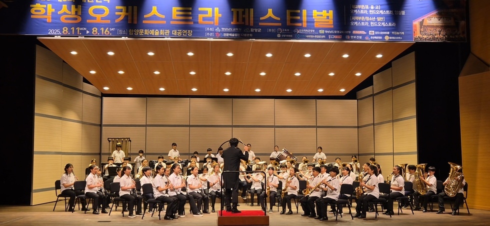 학생들이 함양문화예술회관 대공연장에서 오케스트라 공연을 하고 있으며, 지휘자가 무대에서 연주를 이끌고 있다.