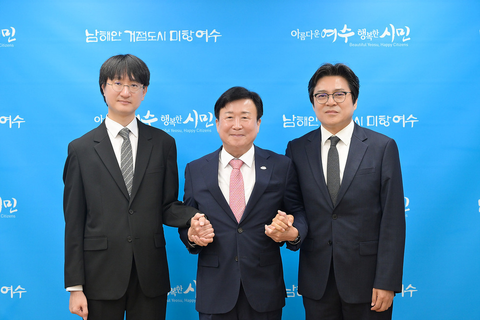 세 명의 남성이 손을 잡고 서 있으며, 배경에는 남해안 거점도시 미항 여수와 아름다운 여수 행복한 시민이라는 문구가 적힌 파란색 배경이 있다.