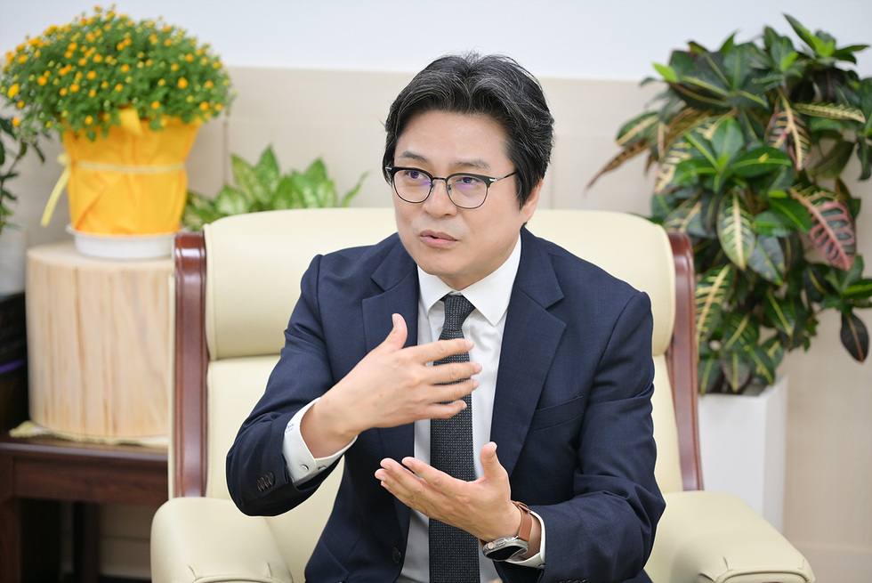 한 남성이 소파에 앉아 손짓을 하며 대화하고 있는 모습이다. 배경에는 식물과 화분이 보인다.