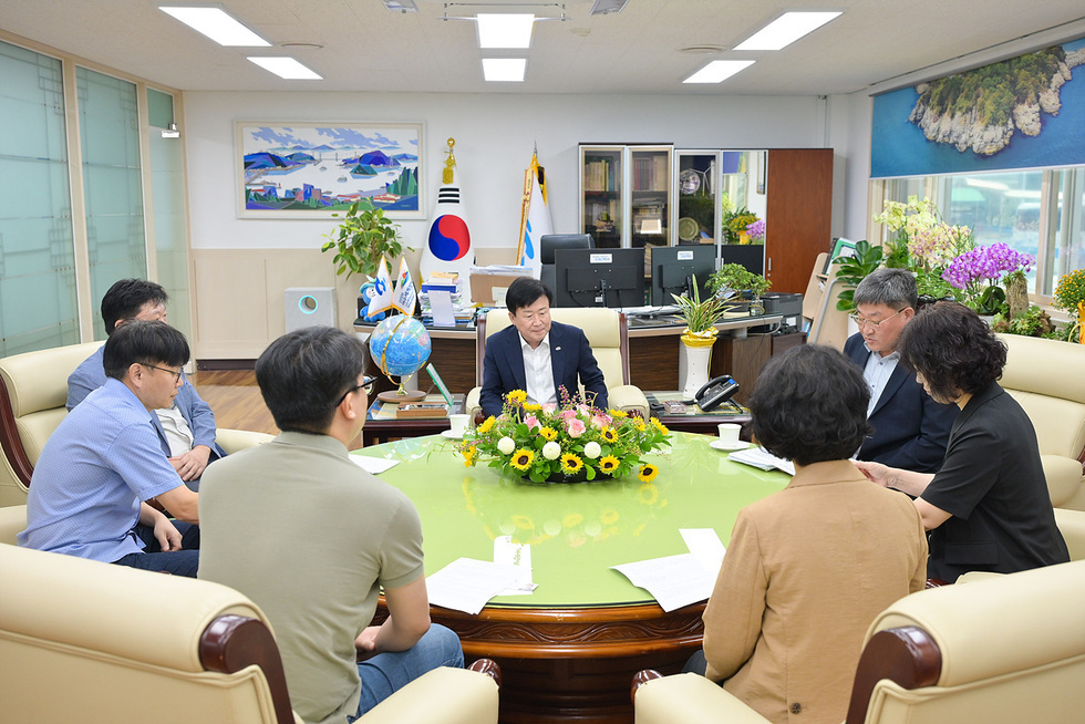회의실에서 여러 사람이 모여 앉아 대화하고 있으며, 중앙에 꽃다발이 놓여 있다. 배경에는 국기와 지구본이 보인다.