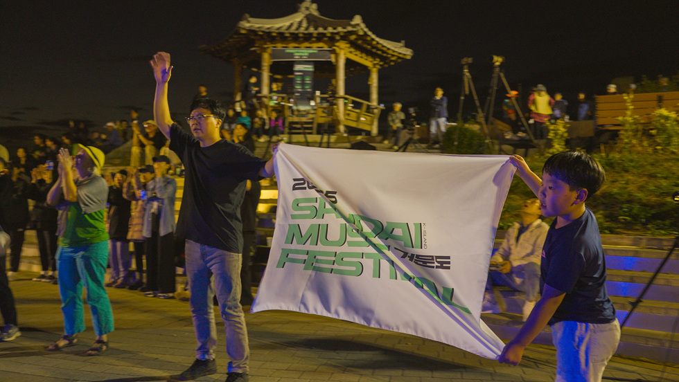 관객들이 2023 산다이 뮤직 페스티벌의 현수막을 들고 응원하는 모습으로, 음악 축제를 즐기는 분위기가 느껴진다.