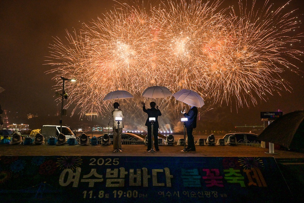 여수밤바다 불꽃축제에서 우산을 든 세 사람이 무대 위에서 불꽃을 배경으로 서 있는 모습. 밤하늘을 가득 메운 황금빛 불꽃이 터지며 화려하게 빛나고 있다. 무대 아래에는 ‘2025 여수밤바다 불꽃축제’ 현수막이 걸려 있다