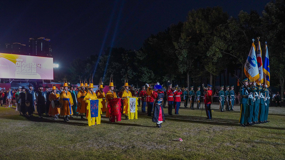 밤이 내려앉은 웅천친수공원에서 열린 여수마칭페스티벌 메인 행사. 전통 복장을 입은 의장대와 군악대, 깃발을 든 참가자들이 나란히 서 있으며 조명이 무대를 밝히고 있다.