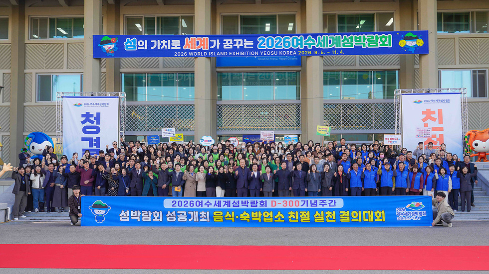 여수시청 앞에서 진행된 2026여수세계섬박람회 D-300 기념주간 결의대회 단체사진. 참가자들이 파란색과 초록색 단체복을 입고 ‘친절 실천 결의’ 현수막 뒤에서 기념촬영을 하고 있다.