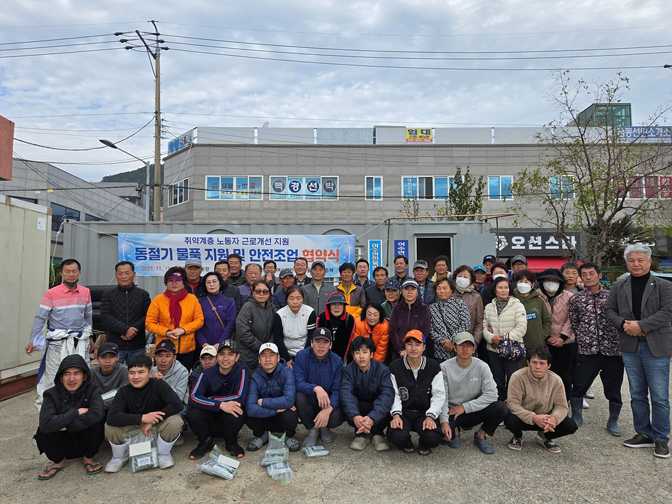 국동항 야외에서 많은 어업종사자들이 단체 기념촬영을 하고 있다. 뒤쪽 컨테이너 건물에는 ‘취약계층 노동자 근로개선 지원 동절기 물품 지원 및 안전조업 협약식(2025. 11. 17./여수시 국동항/여수시노사민정협의회·여수연안복합자율관리공동체)’이라고 적힌 현수막이 걸려 있다.
