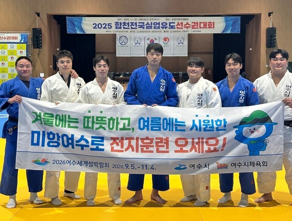 유도 도복을 입은 여수시청 유도팀 선수 8명이 실내 체육관에서 단체로 서서, ‘겨울에는 따뜻하고 여름에는 시원한 미항여수로 전지훈련 오세요!’라고 적힌 홍보 현수막을 펼쳐 들고 있는 모습.