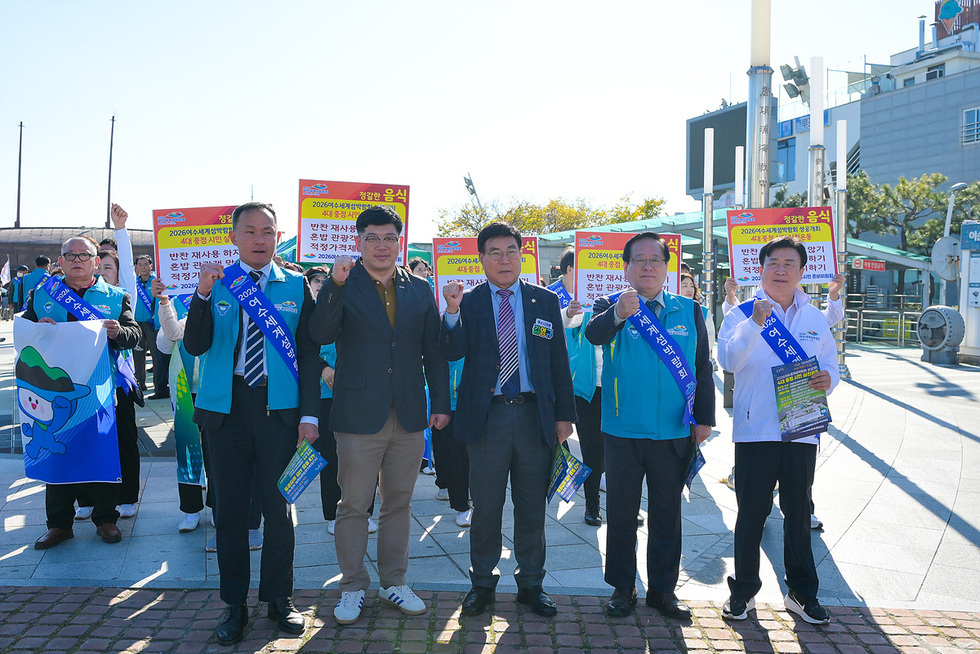 이순신광장 일원에서 2026여수세계섬박람회 성공개최를 위한 시민운동 캠페인을 진행 중인 모습. 파란색 어깨띠를 두른 참가자들이 ‘숙박·교통·음식·청결 4대 실천운동’ 문구가 적힌 피켓을 들고 서 있으며, 앞줄 5명의 인원이 카메라를 향해 주먹을 들어 결의를 표현하고 있다.