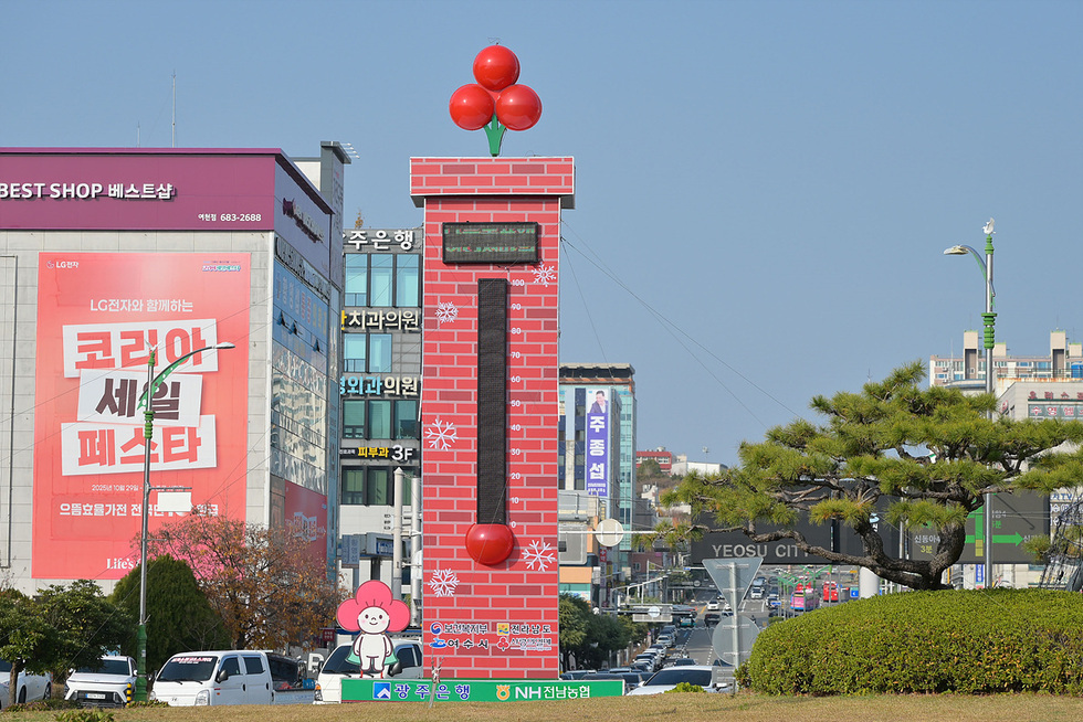 여수시청 앞 로터리 1호 광장에 설치된 붉은 풍선 장식의 사랑의 온도탑이 서 있고 주변 도로와 건물들이 보이며 온도탑 하단에는 여수시청과 NH농협 로고가 표시되어 있다.