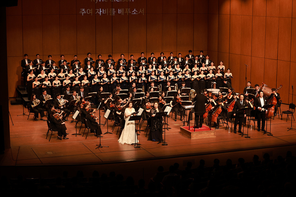 대극장 무대에서 대규모 합창단과 오케스트라가 공연을 펼치고 있다. 수십 명의 합창단원이 계단형 단상에 서서 악보를 들고 노래하고, 무대 앞에는 바이올린·첼로·금관 등 오케스트라가 연주를 이어가고 있다. 중앙에는 흰 드레스를 입은 여성 솔리스트와 검은 드레스를 입은 또 다른 여성 솔리스트가 서서 노래하고 있다