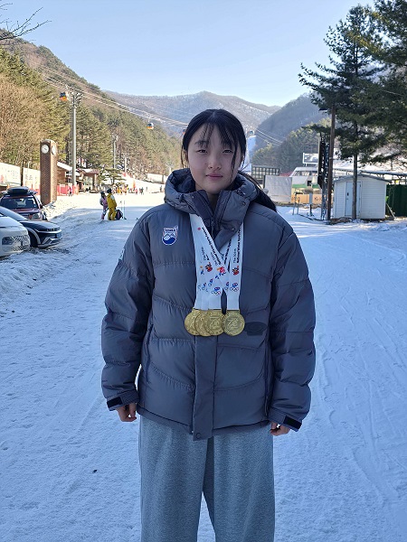 눈 덮인 야외에서 금메달을 목에 걸고 있는 젊은 여성의 모습. 배경에는 산과 스키 리조트가 보인다.