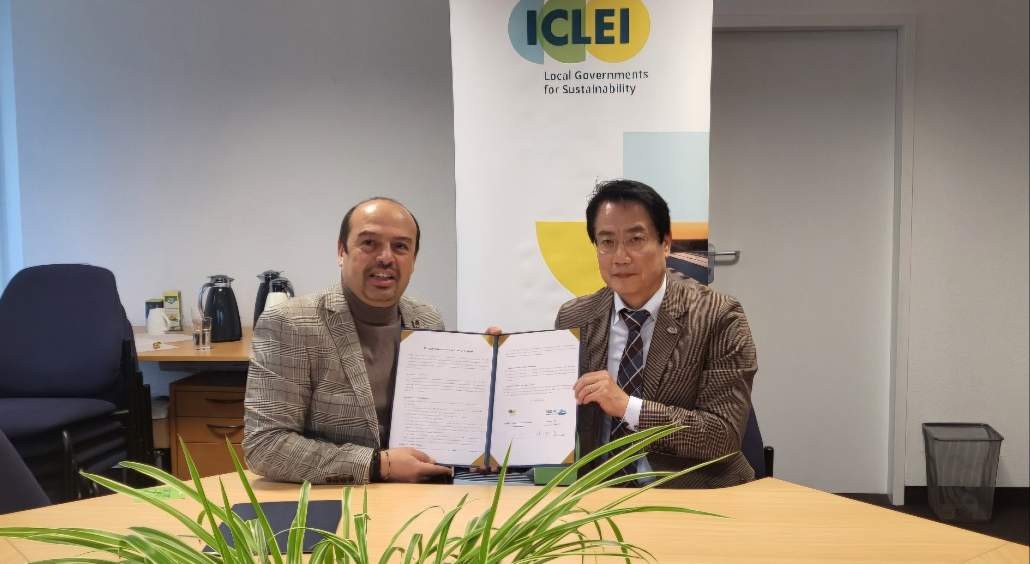 두 남성이 ICLEI 배경 앞에서 문서를 들고 서명하는 장면이다. 이들은 지속 가능성을 위한 지방 정부의 협력을 나타내고 있다.